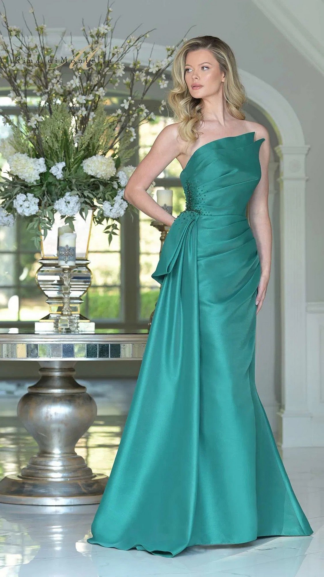 Woman in an emerald Rina Di Montella RD2750 long formal dress stands indoors beside a round table decorated with a large floral arrangement and candles.